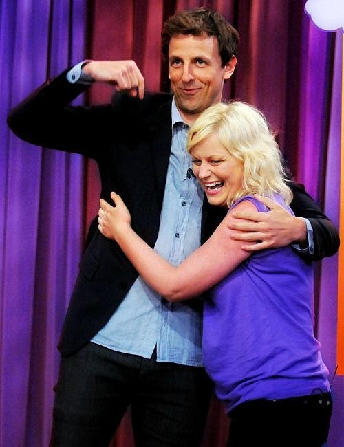Seth Meyers and Amy Poehler hugging with Seth pointing down at her while she laughs.